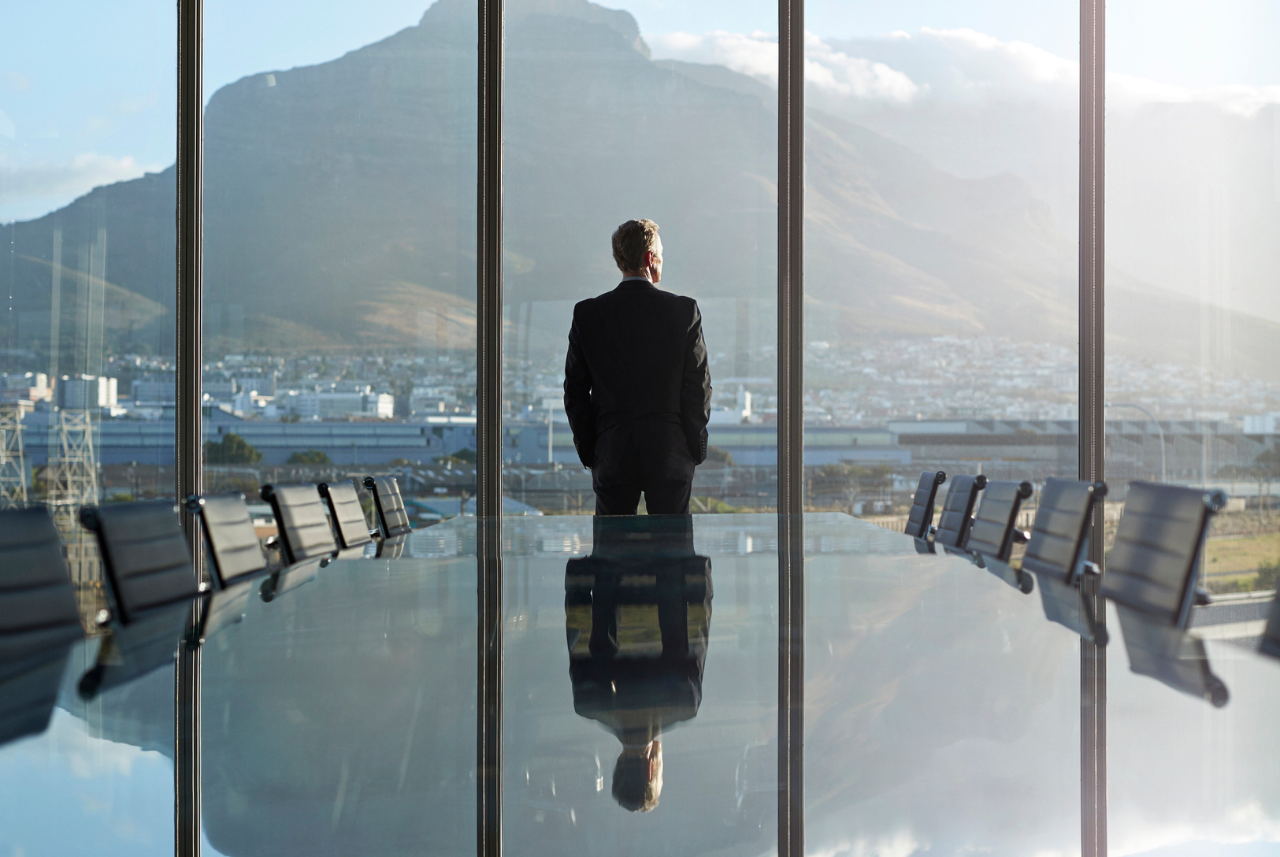 businessman in suit looking out of large conference room window at a town against the backdrop of a mountain